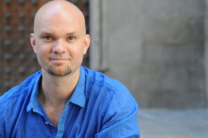 Headshot of a bald person with a beard wearing a blue button-down.