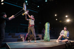 A hawk puppet & puppeteer fight with a man center stage, while another man looks on