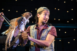 A hawk puppet on a man's arm. The man is wearing a backwards cap, has braids, and a vest