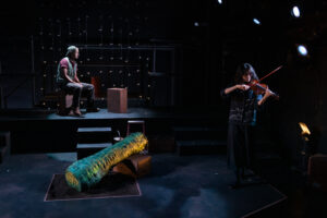 A fem violinist plays toward the right of the stage, while a zucchini puppet sits in the center of the stage
