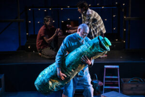 Two brothers paint their nails upstage while at the forefront of the image, a man holds a large zucchini puppet tightly