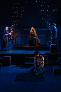 A curtain of string lights is held open by two people: a man in black, and a woman with a light shined on her. She is looking out towards the audience. Behind the curtain is a man holding a large zucchini puppet, and a younger boy sits near soil below