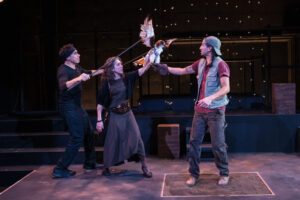 Two puppeteers, a male and female, puppeteer a hawk puppet, while a man in a vest grabs the hawk