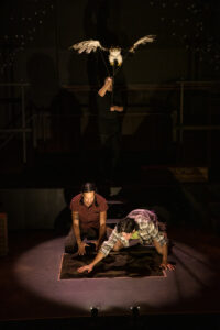 A moody lit scene of two brothers with their hand in soil, and a hawk puppet hovering above them