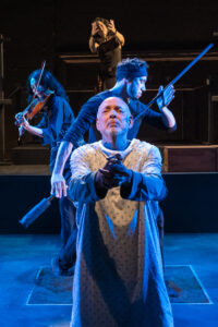 A man at the foreground in a hospital gown with clasped hands, behind him a man holding a long puppetry rod and a violinist face back to back, and then at the very background of the photo, a woman with outstretched hands
