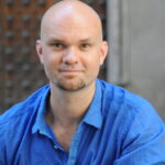 Headshot of a bald person with a beard wearing a blue button-down.