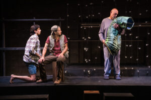 Two brothers sit on a crate and look to their right at their father, holding a large paper zucchini puppet that is wilting.