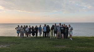 A large group of people stand in front of a lake at sunset