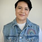 A masc person with a denim vest looks into the camera